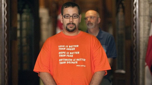 Un homme portant un chandail où l'on peut lire un extrait de la lettre de Jack Layton se recueille devant son cercueil.