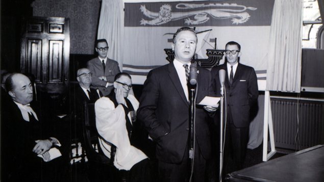 Photo prise � l'Assembl�e l�gislative du Nouveau-Brunswick, le 25 mars 1965, lors du d�voilement public du drapeau du Nouveau-Brunswick.  De gauche � droite, Melbourne M. Hoyt, c.r., greffier du Conseil ex�cutif, le r�v�rend George A. Hatton, pasteur anglican � Fredericton, chapelain de l'Assembl�e l�gislative, l'honorable Louis J. Robichaud, c.r., premier ministre, M. Cyril B. Sherwood, chef de l'opposition � l'Assembl�e l�gislative.  Derri�re, assis, l'honorable Kenneth J. Webber, ministre du Travail et, debout, Robert Pichette, alors chef de cabinet adjoint du premier ministre et concepteur du drapeau.  Collection Archives provinciales du Nouveau-Brunswick. 
