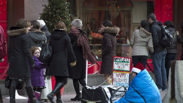 Christmas hoppers walk by a man huddled in a blanket on a Toronto sidewalk under gray Decvember skies.cket