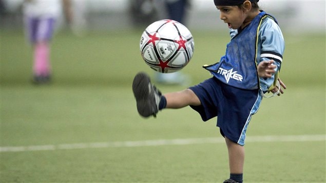 Two-year-old Harshaan Ahluwalis dribbles a soccer ball during a friendly soccer match in solidarity with young players who wear turbans in June 2013 in Montreal. A Quebec turban ban stirred passions earlier this year and people now wonder what will an even broader debate over religious accommodation lead to in Quebec this fall?  Harshaan is wearing a light blue soccer jersey and dark blue shorts. He is balancing a soccer ball about a foot off his right instep with the leg at right angles to his body.
