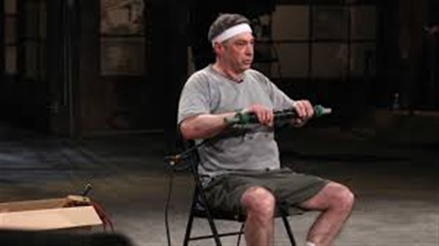 A middle-aged man sits in a sweaty t-shirt and shorts doing arm presses.
