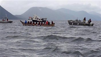 First Nations vessels make their way across gray BC waters with mountains in the background.