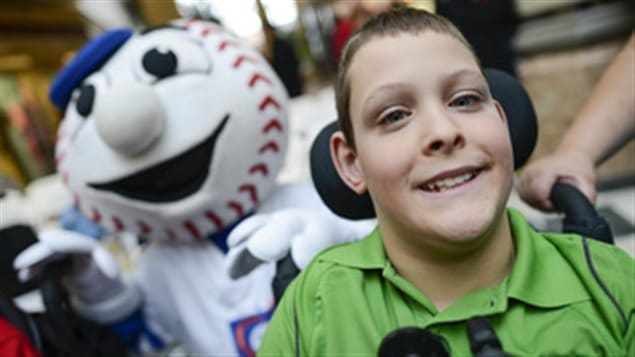 Bryce Durochers in a green shirt with his head leaning back on the head rest of his wheelchair sports a grin while behind him a white baseball mascot with a blue cap and stitches on his baseball head lends support.