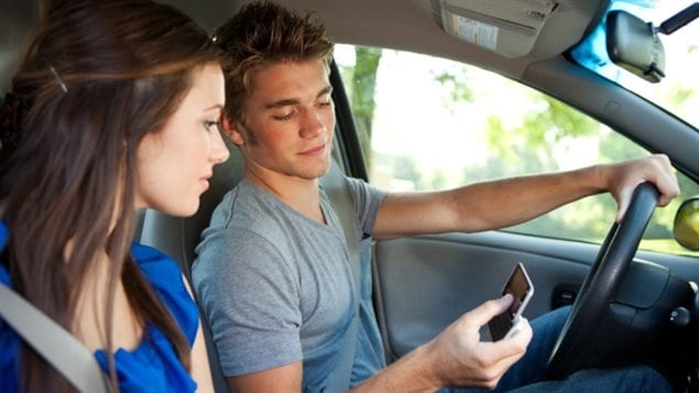Photo shows a teen-aged boy in a grey t-shirt in the driver's seat of a car with his left hand on the wheel holding a smartphone in his right hand. Her is accompanied by a girl in a blue top in the passenger's seat. Both are concentrating on the smartphone.  