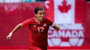 Christine Sinclair will lead the Canadian effort. She is shown in a red Canadian jersey with the number 12 on the chest in white. She is running full force with her left arm forward and her right arm wearing a white sweat band on her right behind her. She has a look of complete determination on her face.