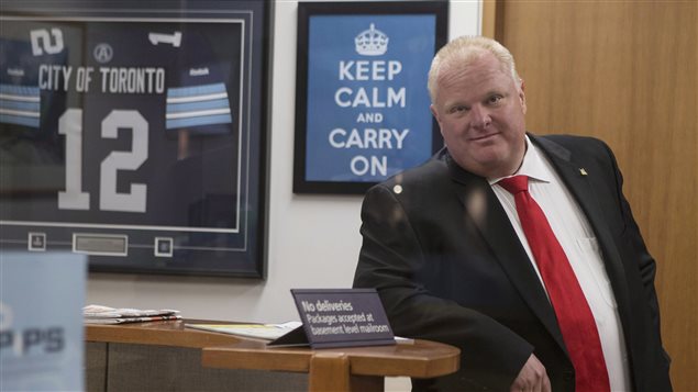 Toronto Mayor Rob Ford waits for an elevator before leaving his office at city hall to take part in a vote on Wednesday. Ford is wearing a brown suit and red tie. He is leaning against a counter. Behind him is a sign that says "keep Calm and Carry On. To the left of that sign his a framed football jersey with the number 12 and "City of Toronto" above the number. 