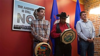 From left to right, Chief Francis Laceese, of the Tl'esqox First Nation, Chief Roger William, of the Xeni Gwet'in First Nation, checking his phone, and Chief Percy Guichon, of the Tsi Deldel First Nation, stand near a poster showing opposition to the Enbridge Northern Gateway Pipeline during a news conference in Vancouver in June. The two chiefs in the left of the picture are holding small, round drums. Behind them are two blue flags against a salmon coloured wall. All three appear to be in the mid to late 40s.