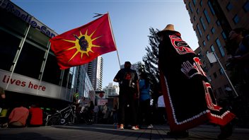 A Mohawk flag is brandished at a rally in June in Vancouver to show opposition to the Enbridge Northern Gateway pipeline. In addition to rallies, First Nations are using legal challenges to fight the building of the pipeline. The flag is red with a yellow sun exuding rays around it. In the middle of the sun is a drawing of a warrior, who has short, black hair and a feather pointing skyward at the back of his head. Walking toward the two people holding the flag is a man in a long, embroidered black coat with orange drawings on it. He is wearing what appears to be a straw hat.