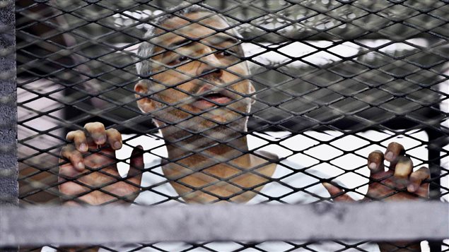 Mohammed Fahmy, the Canadian-Egyptian who was the acting Cairo bureau chief for Al-Jazeera, is shown in a defendant's cage at a courtroom in Cairo, in May. He is waring a white t-shirt and has the fingers of both his hands grasping the wire of the cage.