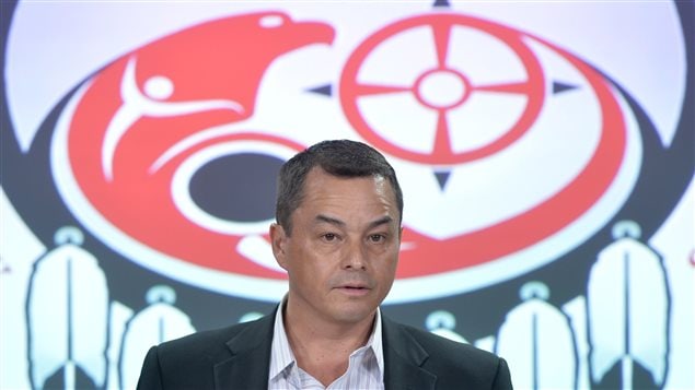Former First Nations chief Shawn Atleo announced his resignation at a news conference in Ottawa earlier this year. The AFN will select a successor in December. Mr. Atleo is seen at a podium without a tie wearing a dark sports jacket and a blue striped dress shirt. Behind him is a large logo featuring what appears to be a red painted thunderbird on a drawing of a drum. A series of four feathers are visible dangling from the painted drum.