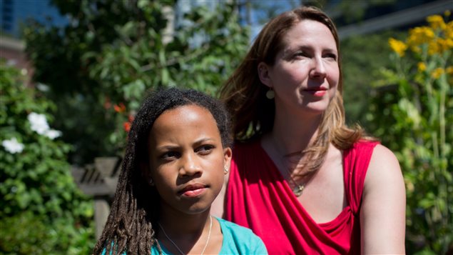 Tracey Wilson and her mother, Michelle Wilson, sat for a photograph in Vancouver on Wednesday after winning a significant equality rights victory. Tracey is a person of colour. We see her from the neck up in the left of the photo. Her mother is a Caucasian with blond hair. She is wearing a red dress exposing her arms and neck. Both have serious expressions on their faces and are looking into the distance. Behind them is greed shrubbery