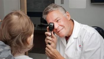 Dr. Robert Koenekoop, who is both an geneticist a ophthalmologist, led a study that offers great hope for many blind people, especially children. He is shown performing an eye examination on a child of bout two or three. Dr. Koenekoop has an instrument help up to his right eye. He is wearing a white lab coat. A handsome man starting to gray, he has a kind face.