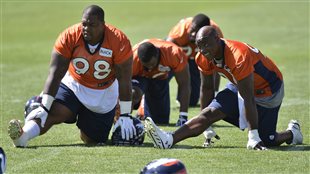 Members of the Denver Broncos stretch at a football camp in June. Research says they should be very careful about how they do it. There are four players in the photo on a perfect green turf. They are wearing orange jerseys and blue shorts. They are on the ground and stretched forward. The man on the right has his right hand touching his right lower leg between his knee and ankle. The man on the left also has his right leg stretched out but is balancing himself by placing  his hands to his side on the ground. The two players pictured behind the two in the forefront are in positions similar to the player on the right--using both hands for balance.