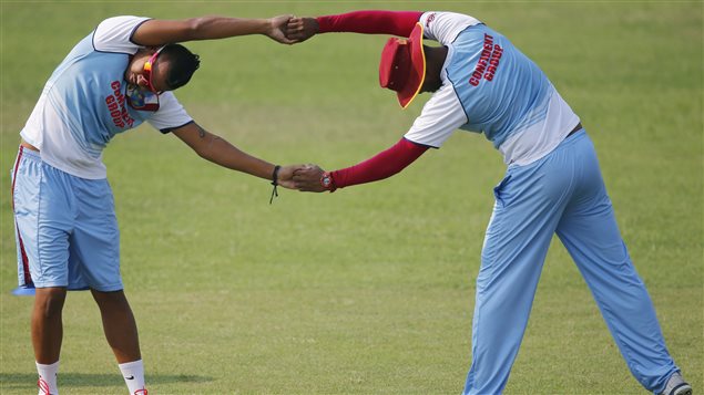 A pair West Indies players perform stretching exercises during a training session ahead of their ICC Twenty20 Cricket World Cup semi-final match against Sri Lanka in Dhaka, Bangladesh in April. Both are wearing mainly teal-coloured outfits. The man on the right is wearing long pants and a red stetson hat. The man on the left is in long, Bermuda-length shorts. Both are standing on a lovely greet pitch. Their arms are locked and they are pulling each to the other. Their jerseys are two-toned. They are mainly teal-coloured with white short sleeves.