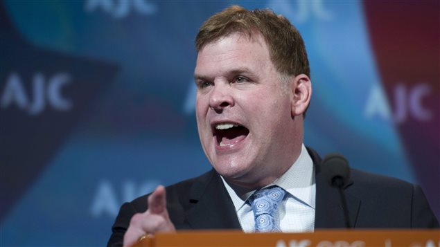 Foreign Minister John Baird gestures as he addresses the American Jewish Committee Global Forum closing plenary in Washington, DC in May. Baird is shown speaking from a lectern with a light blue and black image projected on a wall behind him. The capital letters "AJC" are written on the projections in several places. Mr. Baird is wearing a dark suit, white dress shirt and light blue tie. He is gesturing with his right hand and his mouth is agape as he attempts to make a point.