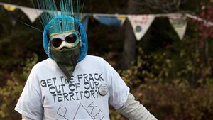 Anti-fracking activists are trying to make their case in many ways. Here, a protester is seen in Rexton, New Brunswick in October, 2013 as police began enforcing an injunction to end an ongoing demonstration. The man is wearing a white t-shirt that says in big black letters GET THE FRACK OUT OF MY TERRITORY. He is shown from just above the waste standing in front of a wooded area. In front of the woods are small pennants bent into various positions by a breeze. On his face, he is wearing dark sun glasses over what appears to be an East Indian light blue mask. Sticking up from the mask are what appear to be long, light blue straw-like hairs that extend to about a foot above his head. Below the mask is a bandana of brown camouflage design. Atop his head is a turquoise-coloured mask of some sort, resembling one a doctor might wear in an operating room.