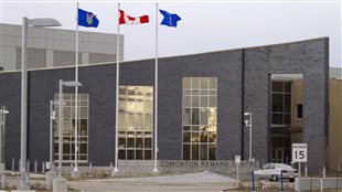 The Edmonton Remand Centre is shown in 2013. More and more people who have not been convicted of a crime are spending more and more time in Canadian jails. The jail has a roof that gradually slopes from right to left. The building is dark grey and has large rectangle windows running from ground level up to about five feet from the roof. The windows are reflecting some sunlight. A grey driveway leads up to the building in front of which (from left to right) fly the Alberta, Canadian and City of Edmonton flags on tall flagpoles. Behind the sloping roof to the left is a higher building. It is institutional grey but lighter grey than the main building. At the front left of the building, a grey street light rises. It is standing on a cement base.