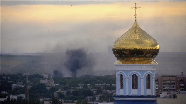 Black smoke billows in Luhansk, Ukraine following a recent mortar attack during fighting between Ukrainian government troops and pro-Russian fighters. In the right foreground of the photo, a golden spire of an Eastern Orthodox church rises. The spire sits atop a light blue and white church structure, dominated by long windows. . In the distance, we see the town which is includes both low-slung singular and row houses and tall, ugly grey apartment buildings. There is an abundance of trees on all streets. On the left in the middle distance, grey-black smoke rises toward a cream-coloured sky mixed with some purple cirrus cloud.