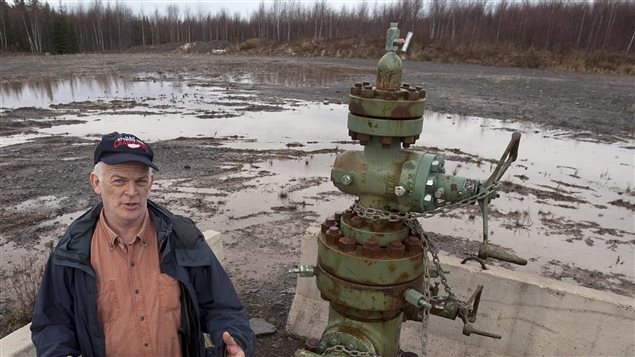 Ken Summers stands next to a wellhead for an exploratory natural gas well near his home not far from Noel, Nova Scotia in December. He is concerned about the impact of fracking on drinking water supplies. He is to the left of the picture, wearing a dark blue baseball hat, open to reveal a salmon coloured shirt. Standing in dark jeans, he is gesturing with cupped hands situated in front of his waste. Beside him is an ugly bit of machinery that raises to about three feet above Mr. Summers's height. It resembles a very tall fire hydrant except it is not red by a rusty grey-green. Behind these two and extending to woods filled with trees devoid of leaves, is area about 200 yards wide of mud mixed with ground water.