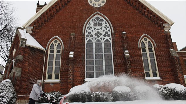 The St. Andrew's United Church in Chatham, Ontario is shown on a winter's day in 2011, two weeks before Kairos, a charity administered the United Church of Canada, was the centre of a contoversy over federal government funding. Kairos says it is now being audited by the Canada Revenue Agency. The photo shows a man pushing a snowblower in front of the church, a high red-brick building with a tall stained glass window centring its front. Two smaller windows flank the large one. Above the tall window, located just below the church's v-shaped roof, is a round window. Shrubs in front of the church are covered with snow. A tall tree to the right of church is bare, perhaps shivering below a grey winter sky.