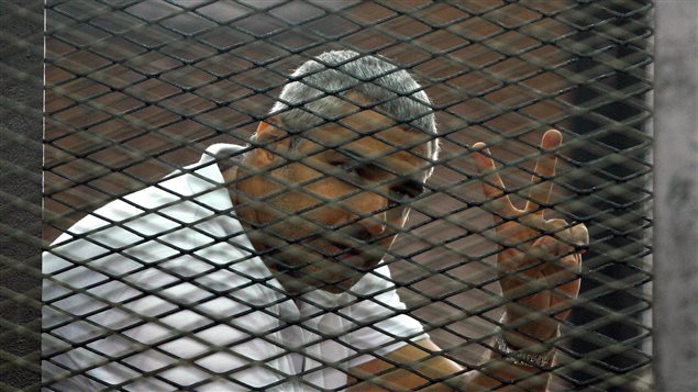 Canadian-Egyptian journalist Mohamed Fahmy gestures from the defendant's cage during a sentencing hearing in a courtroom in Cairo, on June 23. The photo shows Mr. Fahmy from the left, from the chest up, wearing a white polo shirt. He is seen behind mesh like bars and is flashing the peace sign with his left hand. He is slightly hunched over. Mr. Fahmy, a chunky man, has close-cropped grey hair. His face is expressionless.