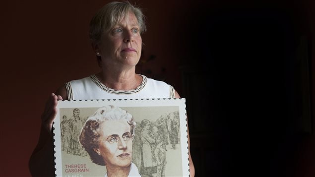 Michele Nadeau, president of the Therese Casgrain Foundation, holds a replica of a stamp depicting her grandmother Therese Casgrain in Montreal on Friday. Ms. Nadeau is an imposing looking woman with grey-blond hair and blue eyes. She is wearing a white dress. The replica of the stamp covers her front, from her waste to her shoulders. The stamp shows her grandmother's face looking outward to her left. She has white and brown curly hair and has a both a kind and formidable face. Behind the image of her face, we see drawings of women and children in working class costumes. To the lower right of the stamp CANADA is written in capital letters of dark brown. To the left, we see the name Therese Casgrain written in red. Directly below that we see the words "LE BIEN COMMUN" and "THE COMMON GOOD" directly below that. Below the row down-, we see the number 32 (the price of the stamp) matching in size the print of the  CANADA previously mentioned on the lower right.