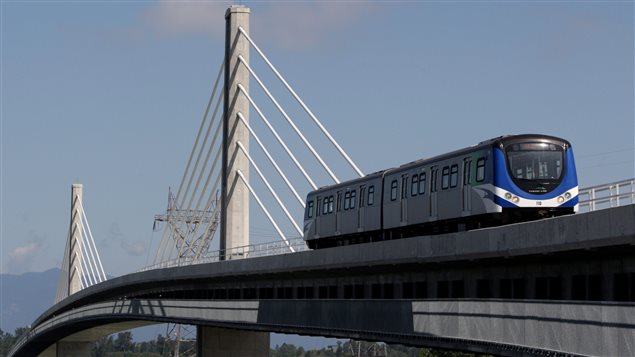 A Canada Line rapid transit train is seen crossing the Fraser River from Vancouver to Richmond. An investigation into problems with the system that stranded thousands of passengers is just getting under way. From below left, we  a two-car silver commuter train, painted mainly blue with white trim at the back end. The train is located at the near right of the photo and is headed toward a tall, grey support stretching perhaps 50 feet into the air. Flowing off the support are white cables that hold the bridge in place. A similar support is located farther along the bridge, which is slightly rounded at its base. Through the bottom of the bridge we see some forest greenery. In the distance we see some dark blue mountains sitting below a powder-blue sky. 