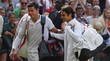 Roger Federer of Switzerland, right, leaves the court with Milos Raonic after the Federer's victory in the Wimbledon semi-finals earlier this summer. Raonic is seeded seventh sixth at the Rogers Cup in Toronto. Federer has a beatific smile on his face and is reaching out with his right hand patting Raonic on his upper back. Raonic, too, is reaching for Federer, but his not yet made contact with his opponent's back. Raonic is poker-faced and looking forward. Raonic has a black equipment bag over his left shoulder and a white one slung his right shoulder. Federer has his bag slung over his left shoulder. One gets the feeling of a match well-played. Spectators--mostly out of focus--watch in the background.