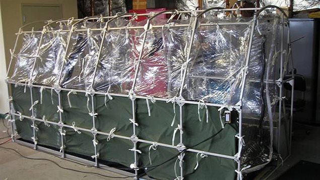 An Aeromedical Biological Containment System which looks like a sealed isolation tent for Ebola air transportation is shown. It is about 10 feet long and shaped by supports extending its entire length. Between the supports is a translucent covering over the upper half. A dark canvas covers the lower half.
