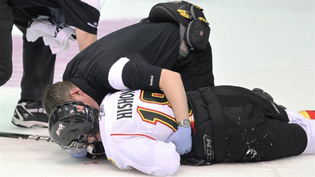 A trainer tends to junior hockey player Joey Hishon (18) of the Owen Sound Attack afterhe was hit in a game against Kootenay Ice at the Hershey Centre in Mississauga, Ontario in 2011. Hishon lost 22 months of his hockey career because of a concussion. The boy is flat on the ice face down. He is wearing a black helmet, white sweater and black hockey pants. A trainer dressed in black has his left hand over Hishon's back and is talking to him.