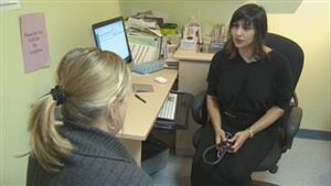 Doctors sharing of more information with patients. Is it a wave of the future? The CMA Journal says it should be. Toward the left of the photo, we see a female doctor, dressed in black and holding a stethoscope, sitting in a chair in a consulting room. She is facing a blond woman, who we see from from the back over her right shoulder. The screen of a computer sits between them while assorted small medical supplies sit on a table to the right of the doctor.