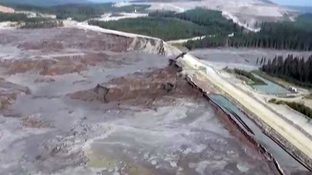 A still photo taken from video provided by the Cariboo Regional District of a helicopter overflight of the stricken area. We see a wide shot of an blue-grey area that appears devastated in the left of the photo. What appears to be a road runs to the area's right. To the right of the road, we see a dark green forest.