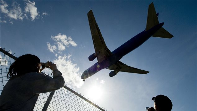 Air Canada flights leaving Montreal will not be bound for Venezuela, at least for now. We see two people taking pictures of a jet silhouetted against an azure sky containing several white whspy clouds. The two people (a woman at the left and a man at the right) are standing behind a chain-link fence looking up at the two-engine aircraft.