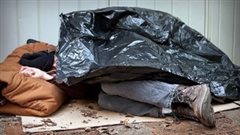 At least 30,000 Canadians are homeless on any given night. We see a man sleeping under a black plastic garbage bag. His feet stick out from underneath while his head rests of what appears to be a sleeping bag.