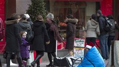 Estimates say 150,000 to 300,000 Canadians experience homelessness in any given year. The photo shows a person huddled under a blue blanked as holiday shoppers walk by in downtown Toronto. A Christmas tree with lights lit is in the background.