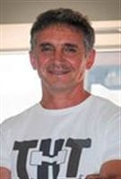 Sam Maniatis is leading workouts for the homeless. We see Mr. Maniatis from the lower chest up. His arms are crossed and he appears to be in very good shape. His has a kind face and salt-and-pepper hair combed back. A smile creases his lips.