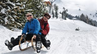 Baby boomers are well aware that things could get tougher as they slide into old age. We see a middle-aged couple (about 55) on a sled on a beautiful, white ski hill with pines and gondola lifts in the background. The man is wearing a blue ski jacket, sun glasses and is beginning to lose his hair, which is greying. The woman behind him is dressed in a red ski jacket and dark scarf. She also is wearing sun glasses and has a full head of dark hair. Both are smiling widely and have their legs pointed forward and up off the ground, exposing the soles of their winter boots.