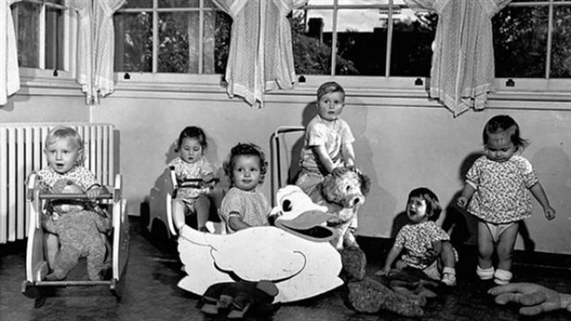 Early baby boomers play with toys in Ottawa, circa 1950. Innocence has been replaced by fear of problems that come with old age. We see a shot of six children aged from about 2 to 3. They are playing in what appears to be a suburban home because through windows above their heads we see trees and a house with a chimney. The child on the left of the picture is sitting in a rocker with a teddy bear in his lap. To his left is a little girl, also in a rocker, who is looking down in a pensive mood. Slightly in front of that child is a little girl sitting in a rocker disguised as a big white duck. To her left and slightly behind is a little blond boy with hair neatly combed looking slightly bewildered by it all. He is sitting on what appears to be a straw ram. To his left are two girls--one sitting, one standing--who must be sisters because they are wearing matching kids dresses.