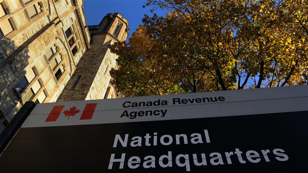 Critics of the Conservative government say charity audits may not start at the CRA headquarters. We see a shot from ground level looking up toward a large government sign that says in white letters on a black background "National Headquarters." Just above that on the same sign we see a drawing of the Canadian flag next to the words "National Revenue Agency." Behind the sign, we look up at the a greystone building and tower. On the right of the photo is a gorgeous maple tree, sitting under a azure sky.