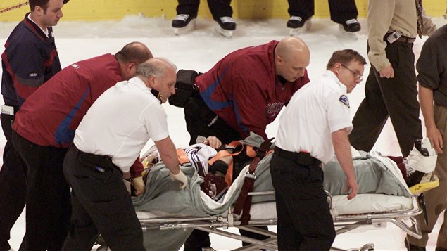 Steve Moore is taken off the ice by medical staff after he was hit by Vancouver Canucks' Todd Bertuzzi on March 8, 2004. We see Moore prone on a stretcher covered in blankets. He is surrounded by medics and support staff rolling the stretcher.