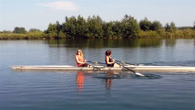 Transgender acceptance took a giant step forward on the weekend. We see two women, one a blond in an orange shirt, the other a brunette dressed in black sitting a two-person racing boat with their oars in water that is dark blue and calm. In the background we see lovely green trees. 
