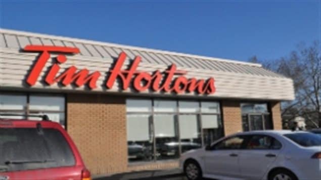 We see a typical Tim Hortons restaurant from the outside. We see two cars parked in front of a modern low-slung one-storey building. The room is medal, the wall between a series of bright windows is red brick. Across the top in orange cursive script are written the words "Tim Hortons."