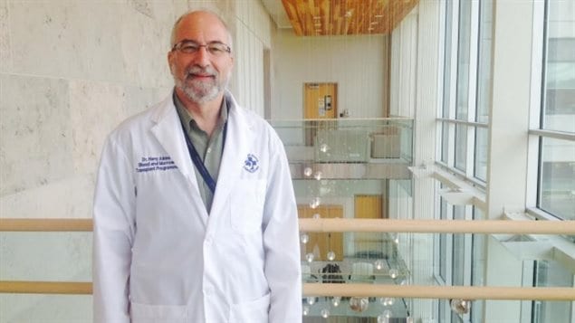 Dr. Harold Atkins had the results of his treatment of Stiff Person Syndrome published in the Journal of the American Medical Association on Monday. We see a balding man, bearded and with glasses, standing an upper floor of the Ottawa Hospital Research Institute. He is wearing a white doctors coat with the blue logo of the institute on right and his name on the left. Through two beige restraining barriers, we look down into an atrium or indoor courtyard topped by many overhanging silver lights. 