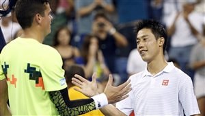 Kei Nishikori, right, shakes hands with Milos Raonic, after their fourth round match at the U.S. Open Open in Tuesday's pre-dawn hours. Both look absolutely spent. We see Nishikori's eyes; they appear glazed, but he has a gentle smile on his face. He is dressed in white. Raonic towers over Nishikori and is wearing a yellow shirt and a black and mesh right arm band and white wrist band. His head is high. It is difficult to read his expression, but he does not appear angry. They are using the clasp handshake, not the traditional handshake.
