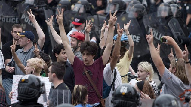 Police encircle and detain a crowd of protesters in Toronto in June, 2010 during the G20 summit. We see a crowd of demonstrators with their arms raised and their hands showing the vee-for-victory sign. They are surrounded by police dressed in riot gear.
