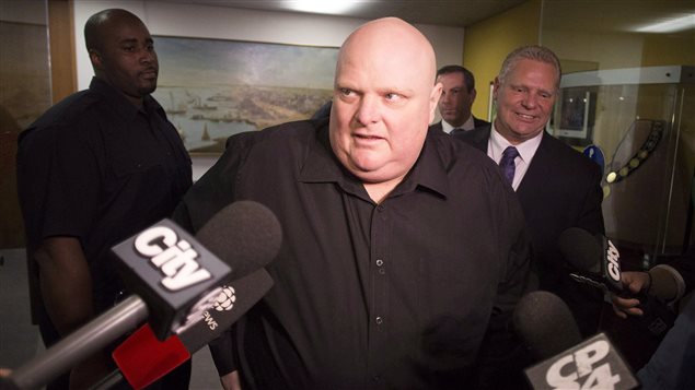 Rob Ford (centre) stands next to his brother Doug Ford (right) outside his office as he scrums with the media before signing bobble heads in Toronto in November. Ford is wearing a dark sports shirt, unbuttoned at the collar. He is a bald, a result of chemotherapy.