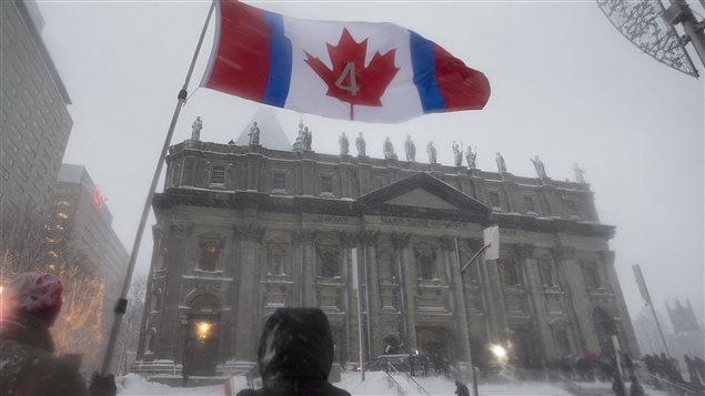 Fans hold a flag in honour of hockey great Jean Beliveau during his funeral in Montreal on Wednesday. The number four in the flag represents the number Beliveau wore as a Montreal Canadien. We see the Canadian flag being held aloft in the foreground. In the background is  Mary, Queen of the World Cathedral. It is dark grey as is the day and the sky.