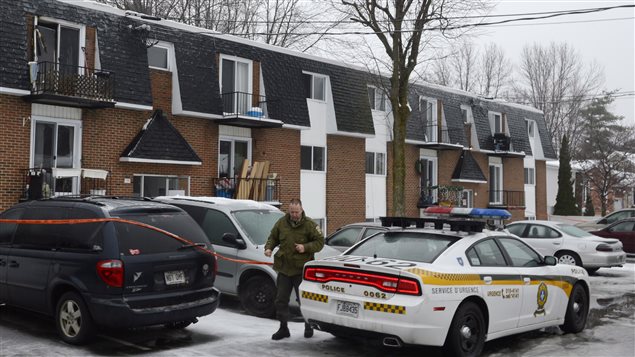 A police officer at the scene of a fire at an apartment building in Les Coteaux, QC., southwest of Montreal early Tuesday. A man and woman and two of their children died. Another child, a eight-year-old boy, is in critical condition at a Montreal hospital. We see a brown-clad police officer walking at the rear of a police car in front of the row of two-storey, brick-fronted apartments. Other cars are parked in front of the building. Grey ice, a grey sky and barren trees accentuate the sombre mood.