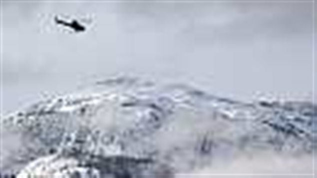 A search and rescue helicopter heads toward a deadly British Columbia avalanche site in 2010. We see a black helicopter in the upper left of the photo. It is flying in a steely grey sky above high, snow-covered mountains