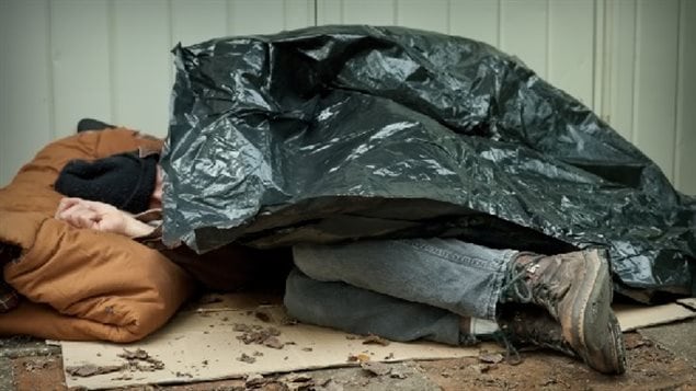 At least 35,000 people in Canada seek a place to sleep in Canada daily. We see the legs and boots of a man sleeping under a garbage bag.